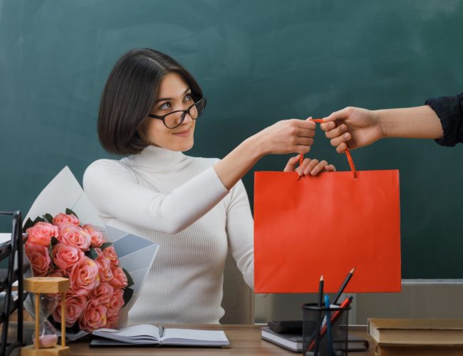 pleased young female teacher received gift bag sitting at desk with school tools in classroom